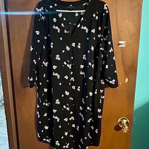 May include: A black dress with a white floral pattern hangs on a white hanger. The dress has long sleeves and a v-neckline. The dress is knee-length and appears to be made of a lightweight fabric.