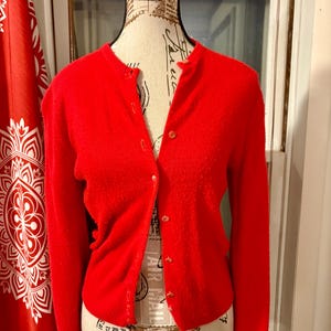 May include: A bright red long-sleeved cardigan sweater featuring a round neckline and a front button-down closure with matching red buttons. The knit garment is displayed on a mannequin, showcasing a fitted silhouette with ribbed cuffs and hem.