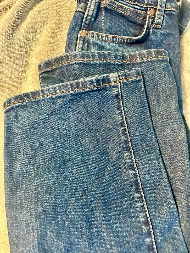 以下が含まれることがあります： 青いデニムジーンズのクローズアップ。ジーンズは折りたたまれており、フロントポケット、ベルトループ、縫い目が見えます。デニムは色あせた外観で、コントラストの効いた黄橙色の糸でステッチが見えます。