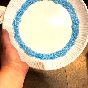 May include: White ceramic plate with a raised, textured rim and a blue, leaf-like border. The plate has a smooth, white center. A hand is holding the plate.