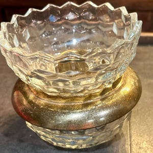 May include: A clear, faceted glass bowl sits atop a gold-toned metal base. The bowl has a decorative, scalloped edge. The base has a wide, rounded shape with a gold finish. The piece is likely a decorative item or a small dish.