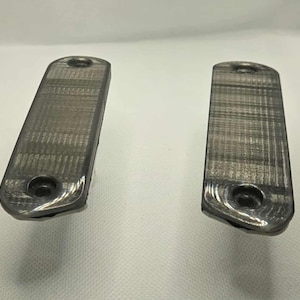 Puede incluir: Dos objetos rectangulares de color gris metálico con extremos redondeados y un agujero en cada extremo. Los objetos tienen una superficie texturizada y parecen ser de metal. Se encuentran sobre un fondo blanco.