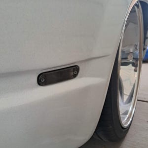 Op de afbeelding: Detailopname van de witte zijkant van een auto, met een donker rechthoekig zijmarkeringslicht dat met schroeven is bevestigd. De afbeelding toont een deel van de band en een chromen velg.