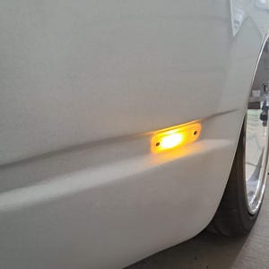 May include: A close-up of a white car's side panel with an illuminated amber side marker light. The light is rectangular and emits a bright glow. A portion of the tire and wheel are visible.