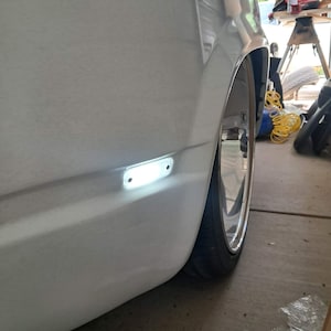 May include: A white car's side panel with a bright, rectangular side marker light. The image shows a chrome wheel and a black tire. The car is parked inside a garage.