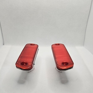 May include: Two rectangular red automotive lights with a textured surface and a circular hole at one end. The lights are positioned side by side against a white background.