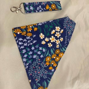 May include: A navy blue dog bandana and matching key fob, both decorated with a floral pattern. The design includes white, yellow, and blue flowers with green foliage. The key fob has a silver clasp.