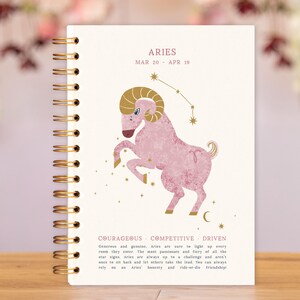 May include: A spiral-bound notebook featuring the Aries zodiac sign. The cover displays a pink ram illustration with gold accents, the word "ARIES," and the dates March 20 - April 19. The notebook also includes text describing Aries traits: courageous, competitive, and driven.