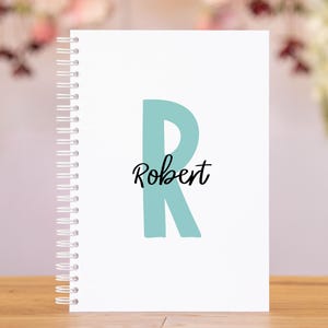May include: A white spiral-bound notebook with a large teal letter "R" and the name "Robert" in black script. The notebook is on a wooden surface, with a blurred floral background.