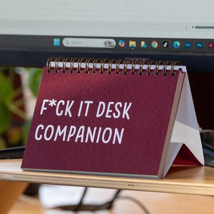 May include: A burgundy desk calendar with the text "F*CK IT DESK COMPANION" in white. The calendar is spiral-bound and stands on a wooden desk, with a computer monitor in the background.