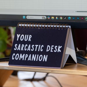May include: A desk calendar with a dark blue cover and white text that reads "YOUR SARCASTIC DESK COMPANION." The calendar is spiral-bound and stands on a wooden desk, with a computer monitor in the background.