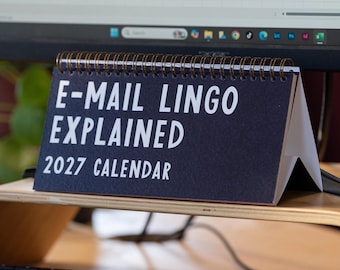 May include: A dark blue desk calendar with white text that reads E-MAIL LINGO EXPLAINED 2027 CALENDAR. The calendar is held together by a bronze-colored metal spiral binding at the top and stands in a triangular shape on a wooden surface.