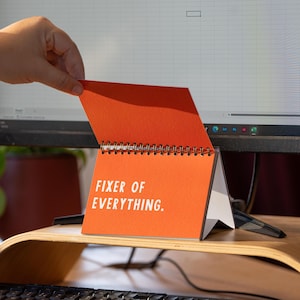 May include: An orange desk calendar with the text "FIXER OF EVERYTHING" in white. The calendar is open, revealing a spiral binding and white pages. The calendar is on a wooden desk stand, with a computer monitor in the background.