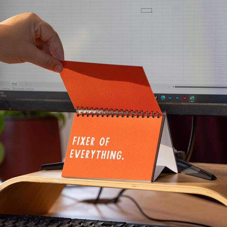 May include: An orange desk calendar with the text "FIXER OF EVERYTHING" in white. The calendar is open, revealing a spiral binding and white pages. The calendar is on a wooden desk stand, with a computer monitor in the background.