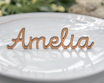 May include: A wooden name cutout that spells "Amelia" in a cursive font. The cutout is light brown and rests on a white plate. The background is blurred, with hints of greenery.