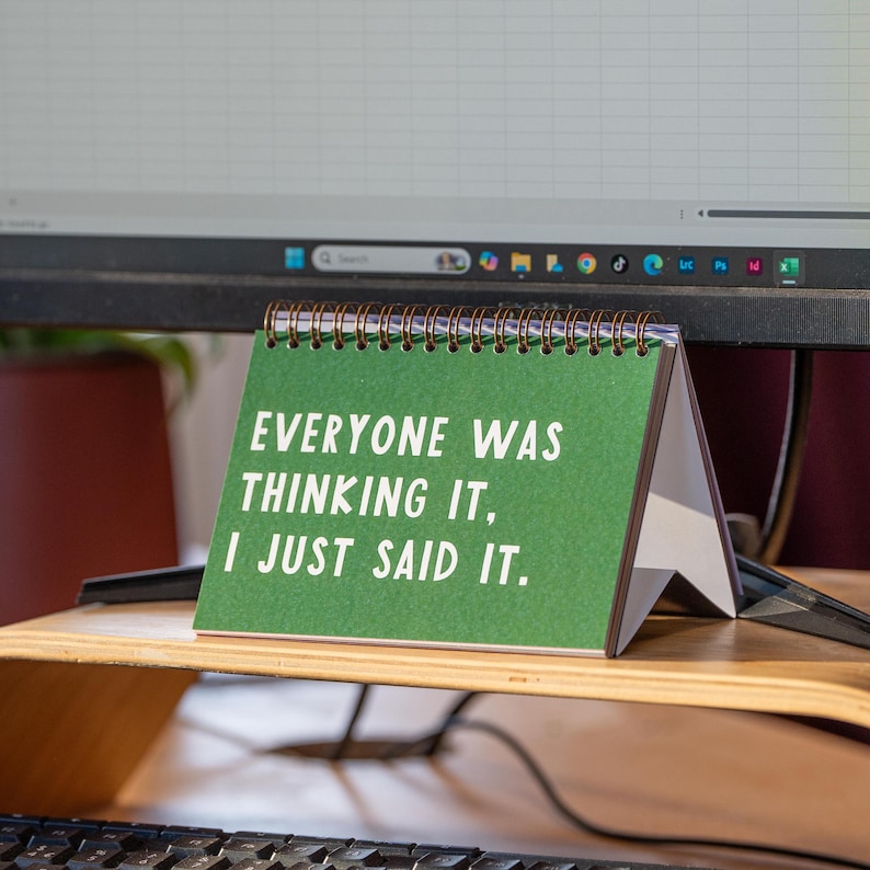 May include: A small desk calendar with a green cover and spiral binding. The calendar displays the text "EVERYONE WAS THINKING IT, I JUST SAID IT" in white. The calendar is on a wooden desk, with a computer monitor in the background.