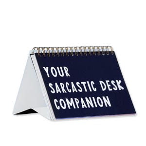 May include: A desk calendar with a navy blue cover and white text that reads "YOUR SARCASTIC DESK COMPANION." The calendar is spiral-bound with a gold-colored wire and has a white stand.