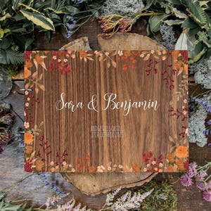 May include: A wooden wedding guest book with the names "Sara & Benjamin" in elegant script. The cover features a floral border with orange, red, and peach-colored flowers and foliage. The date "27th October 2023" is also visible.
