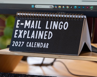 May include: A black desk calendar with white text that reads E-MAIL LINGO EXPLAINED 2027 CALENDAR. The calendar is triangular in shape and features a dark metal spiral binding along the top edge, standing upright on a wooden surface.
