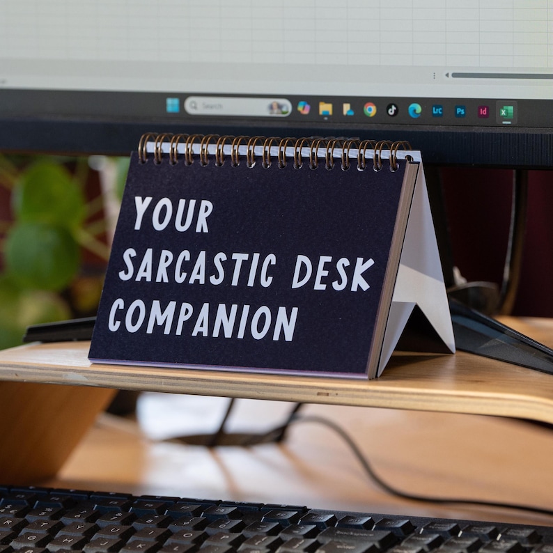 May include: A desk calendar with a dark blue cover and white text that reads "YOUR SARCASTIC DESK COMPANION." The calendar is spiral-bound and stands on a wooden desk, with a computer monitor in the background.