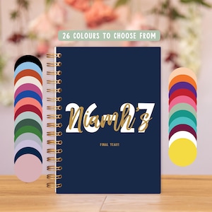 May include: A dark blue spiral-bound planner with gold wire binding. The cover features the white text 26-27, the gold script name Niamh's, and the text FINAL YEAR!!. Two vertical columns of 26 circular color swatches flank the planner.