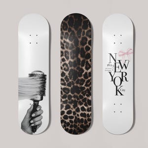 May include: Three skateboards with different designs. One features a black and white image of a hand holding a hairbrush. The center board has a brown and black leopard print. The third board has the text "NEW YORK City" with a pink bow.