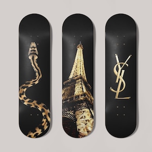 Puede incluir: Tres monopatines negros con diseños dorados. Uno presenta una serpiente, otro la Torre Eiffel y el último el logotipo de YSL. Los monopatines están dispuestos verticalmente sobre un fondo neutro.