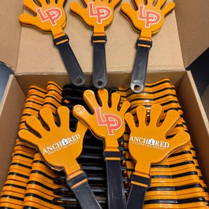 May include: Orange hand-shaped noisemakers with black handles. The hands have the letters "LP" in red and the words "ANCHORED PROPERTIES GROUP" in white. The noisemakers are in a cardboard box.