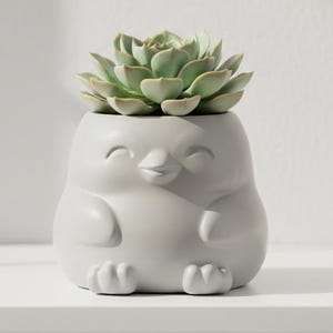 Puede incluir: Una maceta de cerámica gris claro con forma de pingüino, con una cara sonriente y pies pequeños. La maceta contiene una planta suculenta verde. Un artículo decorativo para la decoración del hogar.