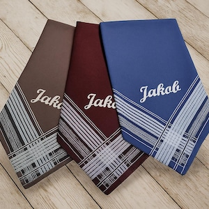 Könnte beinhalten: Drei personalisierte Taschentücher in Braun, Burgunderrot und Blau, jeweils mit dem Namen "Jakob" in weißer Schrift. Die Taschentücher haben ein gestreiftes Design in Weiß und Hellblau.