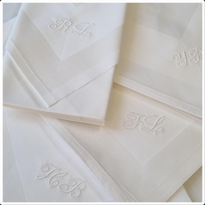 Könnte beinhalten: Ein Stapel weißer Leinen-Servietten mit gestickten Initialen. Jede Serviette zeigt unterschiedliche Initialen in zarter Schrift. Die Servietten haben ein dezentes geometrisches Muster und einen fertigen Rand.