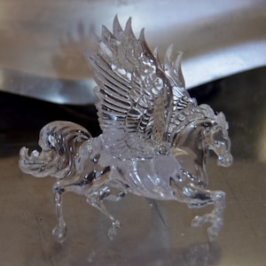 May include: A clear, translucent figurine of a winged horse in a dynamic, mid-gallop pose. The sculpture features detailed, textured feathers on the upright wings, a flowing mane, and a raised tail, all rendered in a smooth, glass-like material.