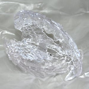 Op de afbeelding: Een helder, doorschijnend ornament van gegoten hars in de vorm van een paar engelenvleugels. Het object heeft een sterk getextureerd, kristallijn oppervlak met gedetailleerde veerstructuren, wat zorgt voor een glanzende en reflecterende uitstraling.