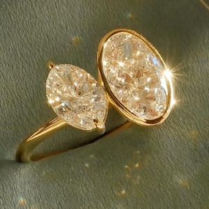 May include: A gold ring featuring two large, sparkling, clear gemstones. One gemstone is oval-shaped, and the other is a marquise cut. The ring is set on a green velvet surface. The text "CHICCHARMSTUDIOUS" is visible.