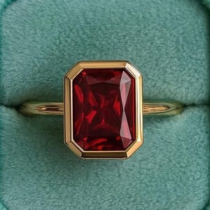 May include: A gold ring with a rectangular, deep red gemstone set in a gold bezel. The ring is displayed in a teal velvet ring box. The text "chiccharmstudious" is at the bottom of the image.
