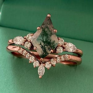 Peut inclure: Un ensemble de bagues en or rose avec une pierre centrale unique en agate mousse et une alliance assortie. L'agate mousse présente des inclusions vertes et blanches. L'ensemble est présenté dans un écrin en velours vert avec le texte "ChicCharmStudious".