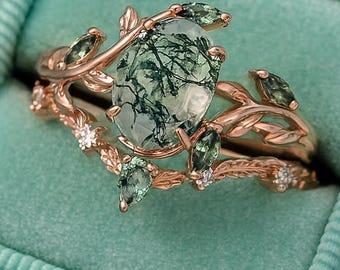May include: A rose gold ring set with a large oval moss agate gemstone, and leaf and vine detailing. The set includes a matching band with small gemstones, presented in a teal velvet ring box. The text "chiccharmstudios" is visible.