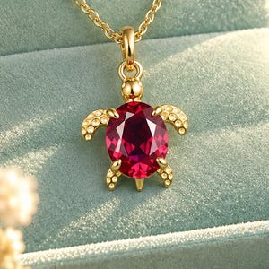 Puede incluir: Un collar de tono dorado con un colgante en forma de tortuga, que presenta una gran piedra preciosa de color rojo intenso con corte ovalado como caparazón y pequeñas piedras transparentes en las aletas. La joya se presenta en un estuche azul.