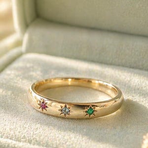 Puede incluir: Un anillo de oro con tres piedras preciosas de colores: rojo, azul y verde. El anillo se muestra en una caja de terciopelo verde claro con el texto "Chiccharmstudious". La caja está abierta y el fondo está suavemente iluminado.