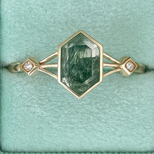 Puede incluir: Anillo de oro con una gran gema verde hexagonal, engastada en un diseño geométrico. El anillo se muestra en un joyero de terciopelo verde claro. Pequeños diamantes flanquean la piedra central. El texto "chiccharmstudious" está en la parte inferior.
