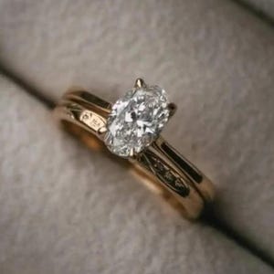 Peut inclure: Une bague de fiançailles en or avec un diamant ovale, présentée dans un écrin gris. La bague présente un anneau fin et une pierre centrale proéminente, offrant une esthétique intemporelle et sophistiquée.