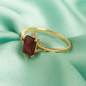 May include: A gold ring featuring a rectangular, deep red gemstone. The ring's band and setting are a bright gold color, with intricate detailing around the gemstone. The ring is set against a soft, teal-colored fabric.