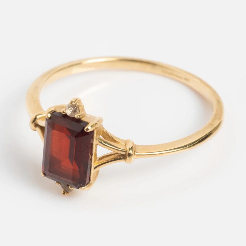 May include: A gold ring featuring a rectangular, deep red gemstone. The ring has a delicate design with a small diamond accent. The band is a simple, polished gold. This piece is a classic jewelry item.