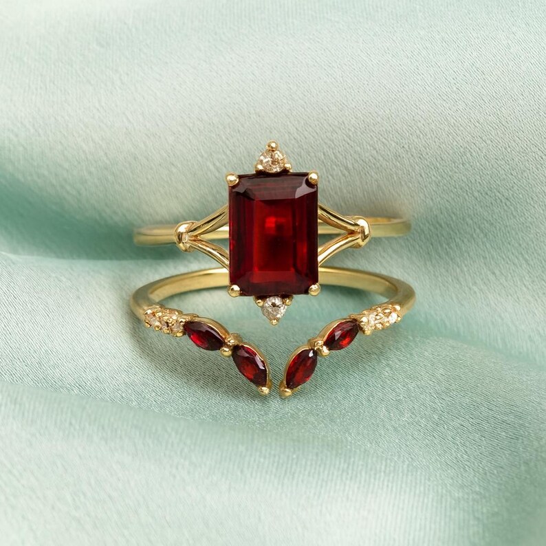May include: Two-piece gold-toned ring set. The top ring features a large, rectangular, deep red gemstone. The second ring is curved, with smaller red gemstones and small white stones. The rings are set against a light blue background.