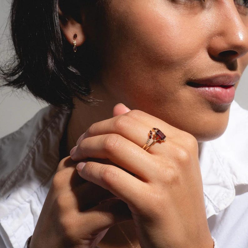 May include: A gold ring with a rectangular, dark red gemstone and smaller accent stones. The ring is on a person's finger, and a matching earring is visible. The jewelry is set against a neutral background.
