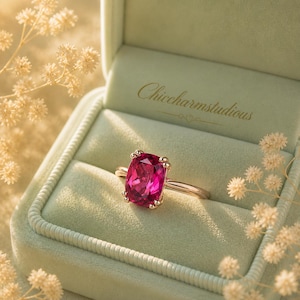 Pode incluir: Um anel de ouro rosa com uma grande pedra preciosa magenta com corte almofada. O anel é apresentado numa caixa de anel de veludo verde claro. A caixa tem o texto "Chiccharmstudious" em letras douradas.