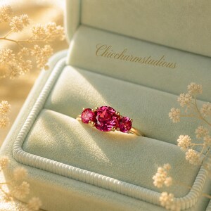 May include: A gold ring featuring three bright pink gemstones, presented in a light blue velvet ring box. The ring is the focal point, with the inscription "Chiccharmstudious" visible inside the box.