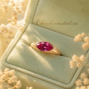 May include: A gold ring with a large, oval-shaped, bright pink gemstone set in a bezel. The ring is displayed in a light green velvet ring box. The text "Chiccharmstudious" is visible on the box.