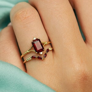 May include: Two gold rings are displayed, one with a rectangular, deep red gemstone and small white stones, and the other with a curved design featuring smaller red gems. The rings are on a finger.