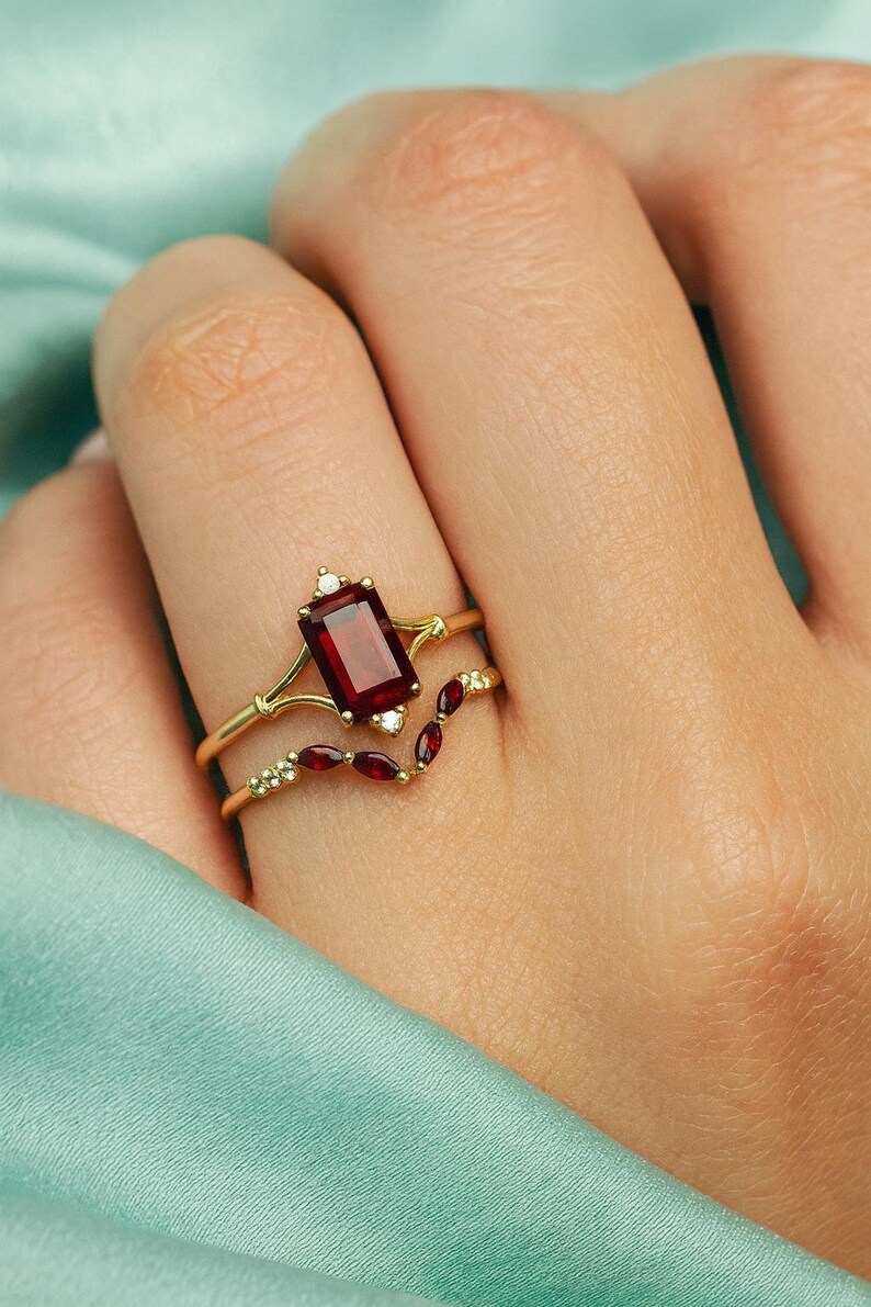 May include: Two gold rings are displayed, one with a rectangular, deep red gemstone and small white stones, and the other with a curved design featuring smaller red gems. The rings are on a finger.